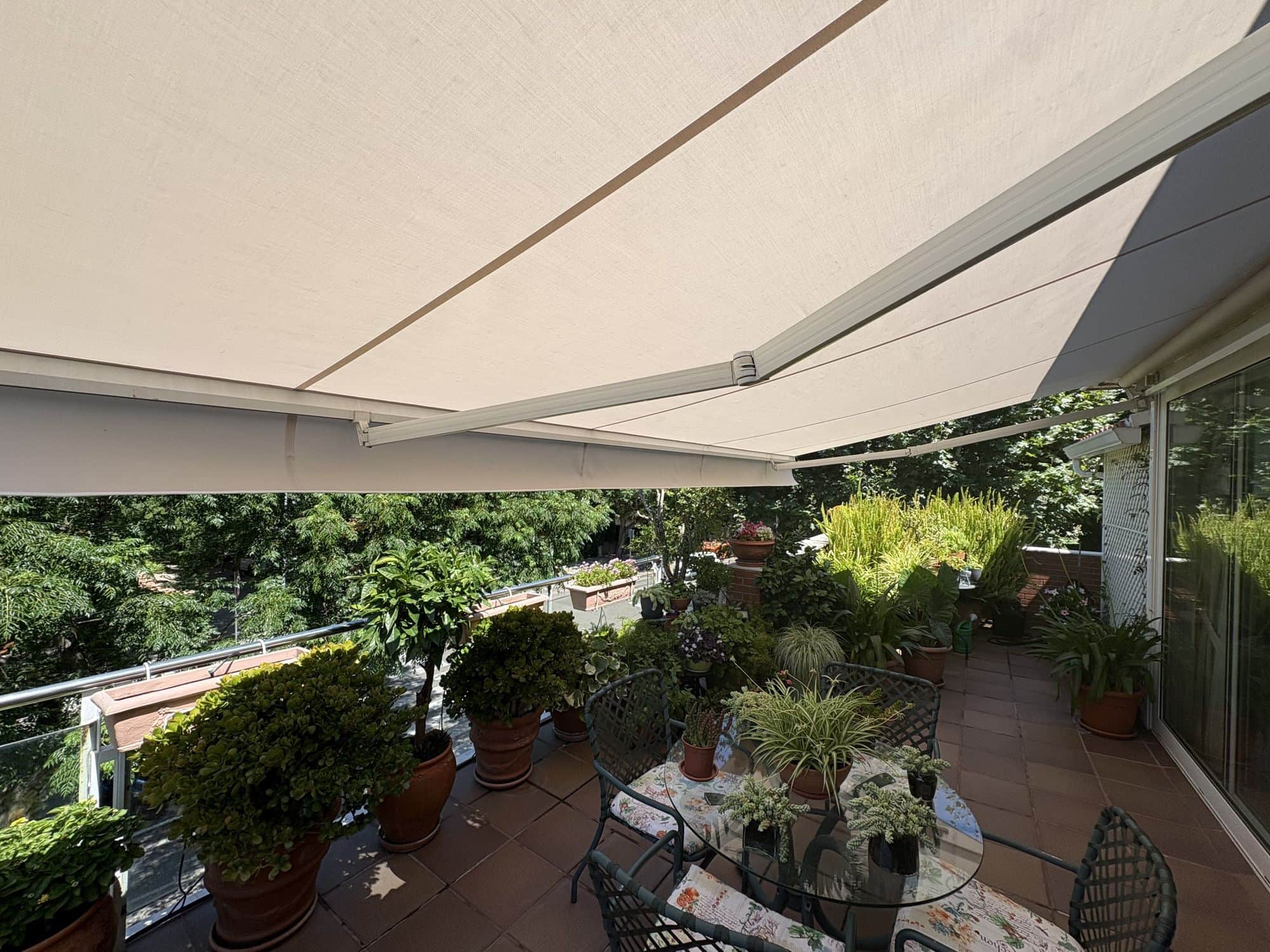 Toldos de brazos invisibles con lona marfil, vistos desde debajo, dando sombra a una terraza con plantas y una mesa