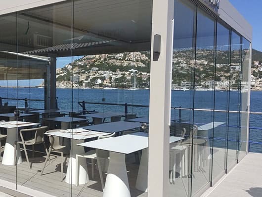 Pérgola bioclimática SEESKY-BIO con cerramiento de vidrio sin perfiles verticales en una terraza junto al mar (imagen del fabricante)