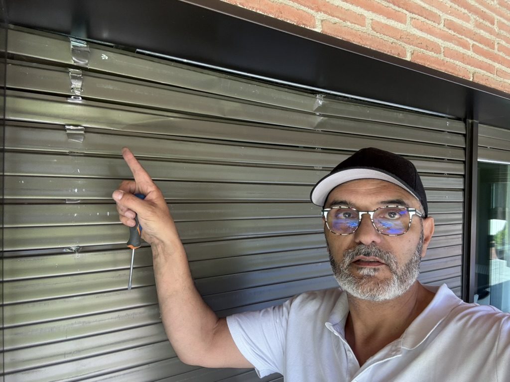 Sergi, destornillador en mano, señalando las lamas de una persiana gris oscura instalada en una fachada de ladrillo